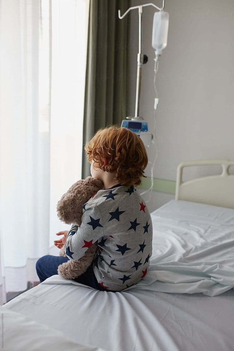 Niño sentado en la cama de una habitación de hospital, abrazando un peluche y mirando hacia la ventana