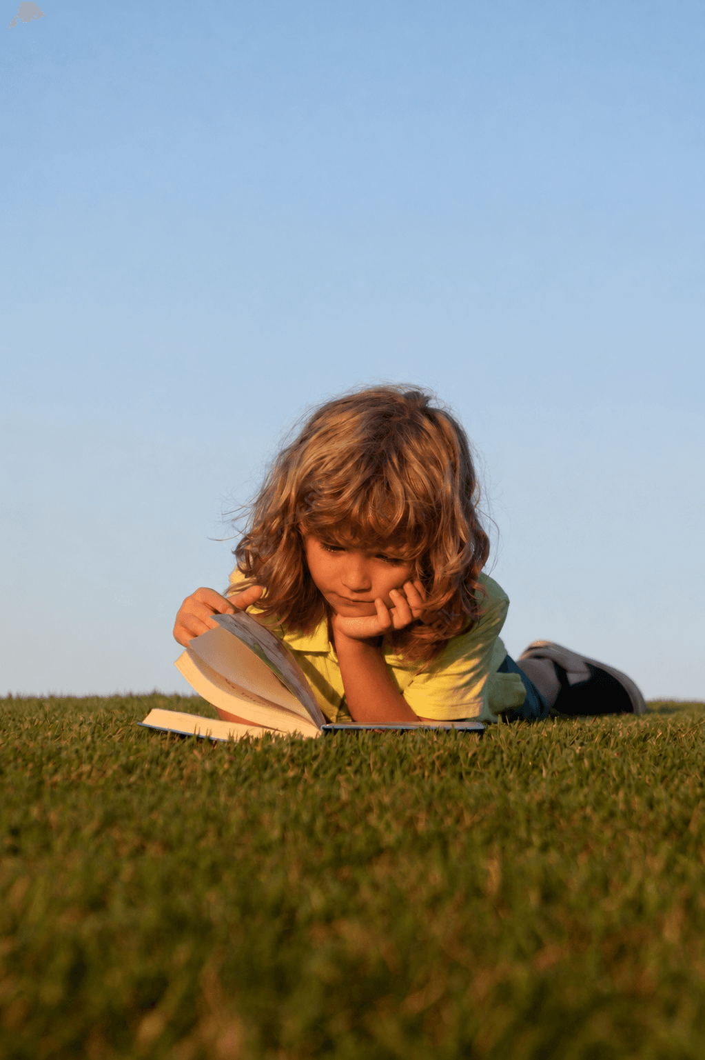 Niño leyendo un libro tendido en el pasto, con el cielo despejado de fondo